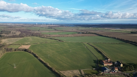 Aerial view of fields and woodland with the sea on the horizon. There are farm buildings in the bottom right corner.