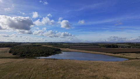 The image depicts a scenic landscape featuring open fields with a small body of water in the foreground. The midground includes a mix of fields and wooded areas. The sky is predominantly clear with scattered clouds, suggesting a sunny day. The horizon stretches to distant hills or low mountains under a blue sky.