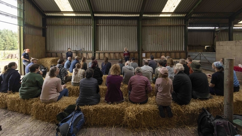 A large group of people sit on three rows of hay bales in a barn listening to a talk for Great North Bog Day 2024