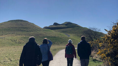 Four people walking away from camera towards Northumberlandia with blue skies above.