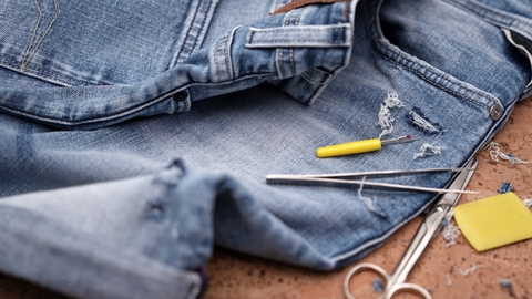 A pair of blue jeans with a hole lie next to sewing equipment including scissors, tweezers and a stitch picker.