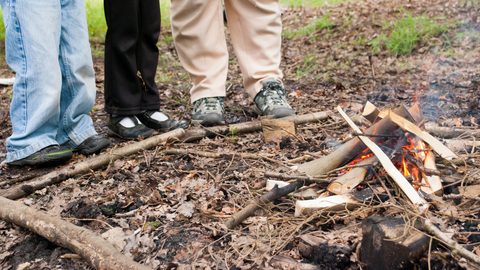 The image shows the lower portions of three people standing around a small campfire.