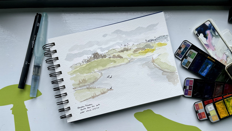 The image shows a sketchbook lying open with a watercolor painting of a landscape, possibly a lake or river scene with greenery and a few birds. Beside the sketchbook are a pen, a water brush, and a palette of watercolor paints. In the background, there's a green silhouette of a snail and a frog on a white surface. The handwriting in the sketchbook indicates the painting is of "Hauxley Discovery Centre."