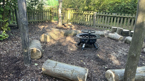Outdoor fire pit area with log seating arranged in a circle, surrounded by a wooden fence and trees.