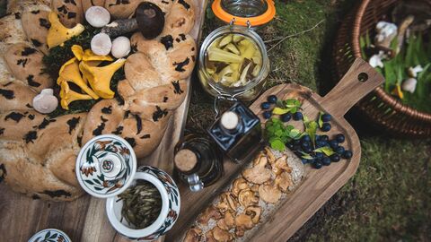 A rustic outdoor food spread displayed on wooden boards. There is a round loaf of bread with dark herbs baked into the crust, surrounded by assorted wild mushrooms. Nearby are jars of preserved vegetables, bottles of dark liquid (possibly oils or syrups), a long slice of bread topped with cooked mushrooms, and small piles of fresh berries and greens. Everything is arranged on a mossy forest surface.