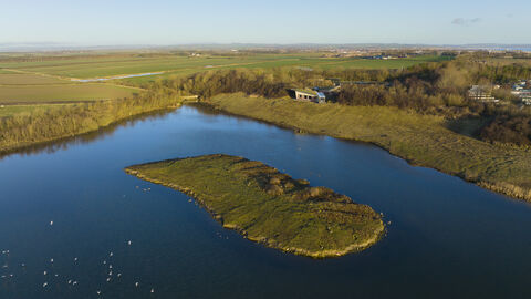 Aerial view of a blue lake with a small grassy island, surrounded by wetlands, woodland, and open farmland under a clear sky.