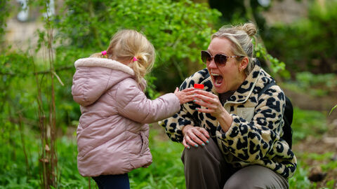 A young child in a pink jacket hands a small red container to an adult wearing sunglasses and a patterned jacket, both standing in a lush green garden.