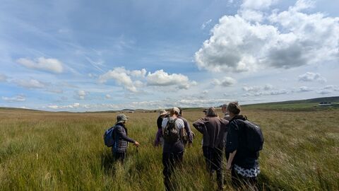 Group of people with backpacks walking through tall grass in an open countryside landscape under a wide blue sky with scattered clouds.