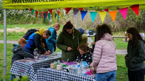 People and children at an outdoor craft stall under a bright “Northumberland Wildlife Trust” gazebo, using colourful materials and tools at a table decorated with bunting.