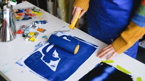 Person using a brayer to roll blue ink onto a printmaking plate on a cluttered table with paint tubes and brushes.