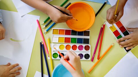 Several hands painting with watercolor sets and brushes around a table covered with colored pencils, paper, and bowls of water.