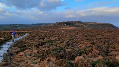 A person with a backpack walks along a stone path through open moorland toward a rocky hill beneath a wide blue sky with large clouds.