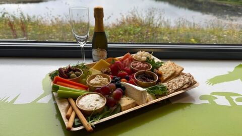 Charcuterie board with cheese, fruit, crackers and a bottle of prosecco on a windowsill overlooking a tranquil lake and greenery.