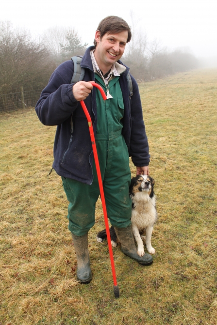 A smiling man in green overalls and wellies standing on grassy field in mist with a dog sat next to him, whilst holding a red shepherds crook. 