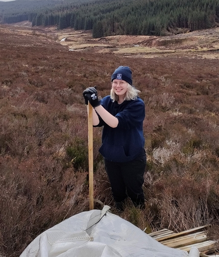 Blonde-haired woman standing on a nature reserve, wearing a blue beanie and work gloves.