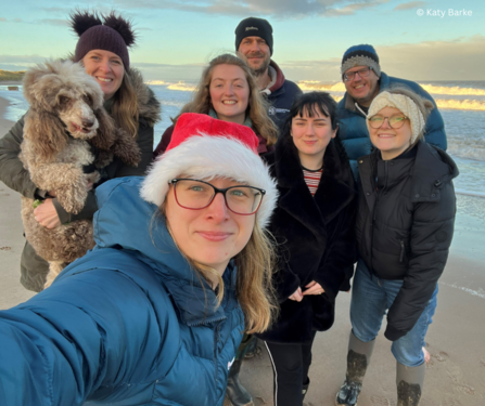 A group selfie photo of staff on a beach. A woman is in the foreground taking the selfie, wearing a Santa hat. Behind her is six other members of staff, one of which is carrying a dog. Everyone is wearing winter clothes and smiling.
