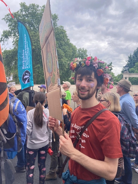 Caucasian man with dark hair and beard, wearing a crown of flowers and a red t-shirt holding a protest sign. Others march behind.
