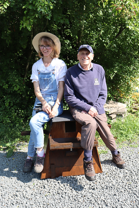 A man and woman sitting outside on a barn owl box in front of bushes.