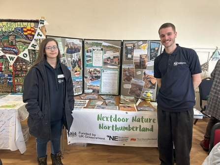 Two staff members in an indoor hall standing in front of a table of information leaflets and flyers.