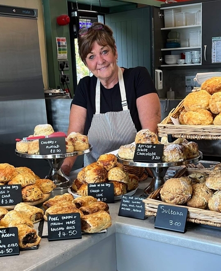 The picture shows a smiling woman standing behind a counter laden with scones and sausage rolls. She is wearing a black t-shirt and a striped apron. The food is displayed on tiered stands and in baskets, with small signs indicating their flavours and prices.