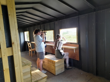 A small group of young people with binoculars sitting and standing in a dark bird hide looking out of the bright windows.