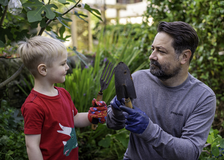 Man and young boy, looking at each other with small garden tools in their hands. Image by Evie and Tom Photography.