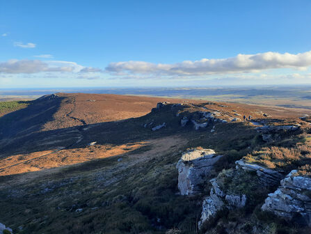 A scenic view of a hilly landscape under a partly cloudy sky. The foreground features rocky outcrops and vegetation, leading to a wide, undulating terrain bathed in sunlight. In the distance, a flatter horizon is visible under the blue sky with scattered clouds.