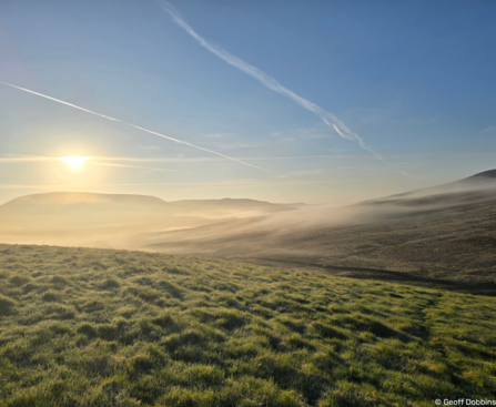 Vast green, grassy landscape with rolling hills. Fog is settled on the ground in the distance and the sun is rising above one of the hills to the left of the image. The sky is clear apart from two airplane trails.