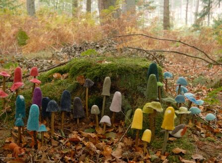 The image shows numerous colourful, felted mushrooms are arranged around the base of a moss-covered tree stump. The mushrooms vary in colour, ranging from reds and purples to blues, greens, and yellows, creating a vibrant contrast against the natural backdrop of brown leaves and green moss. A small, felted bird is nestled among the mushrooms at the bottom right. In the background, there are trees and foliage typical of a forest, with a soft, diffused light adding to the enchanting atmosphere.