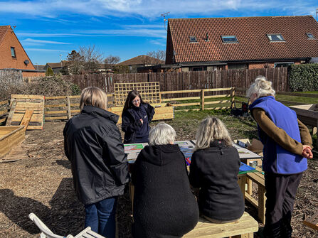 A group of people sit and stand around an outdoor table in a community garden, looking at materials laid out in front of them. Wooden planters, fencing, and nearby houses are visible under a bright blue sky.