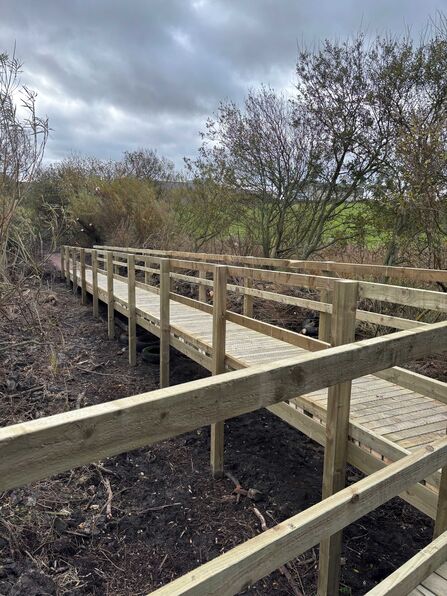 A long wooden boardwalk with handrails runs through sparse vegetation under a cloudy sky.