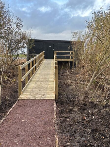 A wooden ramp with handrails leads through sparse trees to a small black wooden building under a cloudy sky.