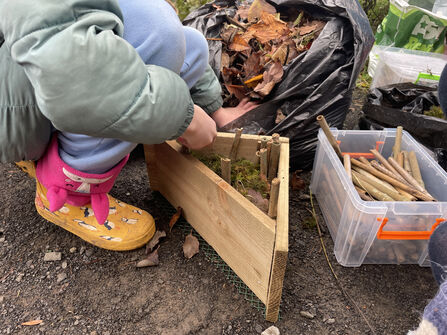 A child wearing a green coat and yellow boots assembles a small wooden bug house on the ground, using moss, leaves, and sticks. Bags of leaves and a container of cut twigs sit nearby.