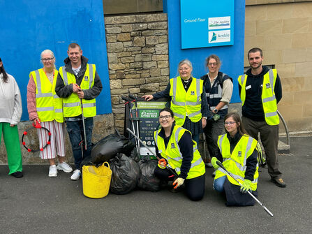 A group of people wearing high‑visibility vests pose together outdoors after a community litter pick, standing beside bags of collected rubbish and holding litter‑picking tools. A blue building wall and signage are in the background.