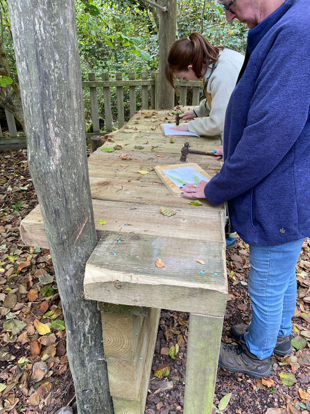 Two people stand at a large rustic wooden table in a wooded outdoor area.