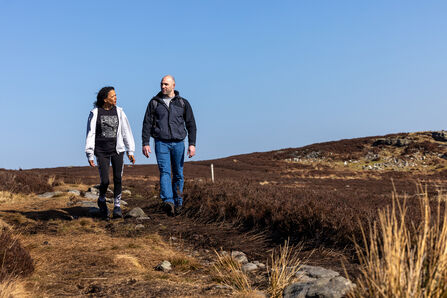 Two people walk along a narrow path through open moorland with low vegetation, under a clear blue sky.