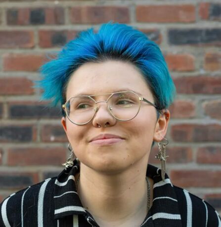 Person with short bright blue hair, round glasses, and dangling earrings, standing in front of a brick wall.