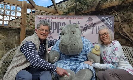 Two adults sit on a bench beside a large plush hippo mascot holding a small toy duck, with a charity event banner behind them in an outdoor garden setting.