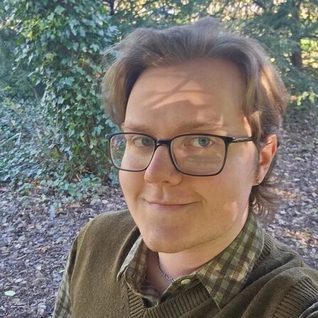 Person wearing glasses takes a selfie on a leafy woodland path, with trees and ivy in the background.