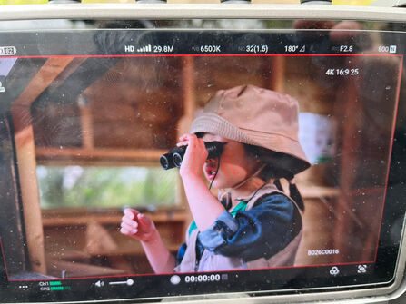 A camera monitor showing a child wearing a bucket hat looking through binoculars indoors, with video recording settings visible on screen.