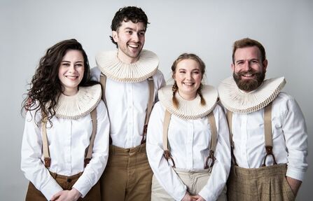 Four people standing side by side in a studio, wearing white shirts, suspenders, and large ruffled collars, with a plain light background.