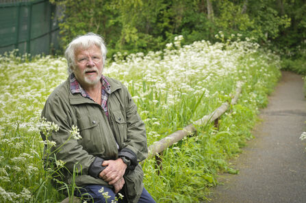 Bill Oddie OBE sitting next to long grass. Image by Chris Taylor Photography.
