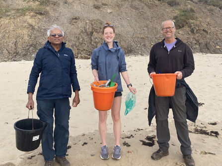 Three people standing on a sandy beach in front of a rocky cliff, each holding collection buckets and beach-sampling equipment during a coastal clean-up.