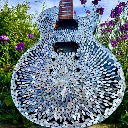 Decorative electric guitar covered in shimmering shell mosaic, standing among purple flowers and green garden foliage under a blue sky.