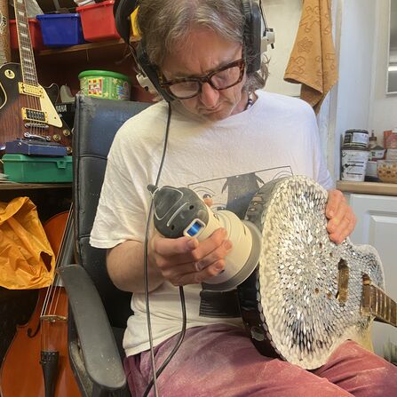 Person wearing protective headphones uses a power polisher on a shell-mosaic guitar in a workshop, with other stringed instruments visible in the background.
