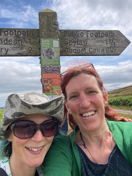 Two people pose beside a wooden public footpath signpost in open countryside, with a partly cloudy sky and rolling hills in the background.