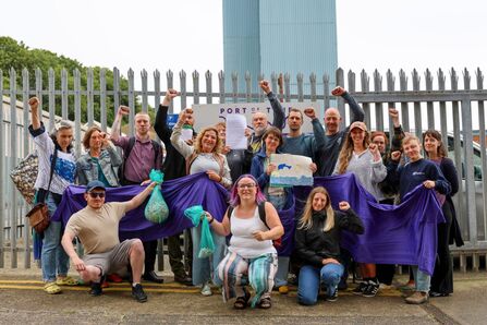 Group of volunteers standing outside a fenced industrial site, holding a large purple banner and bags of collected plastic pellets, with several people raising their fists in support of an environmental clean-up campaign.
