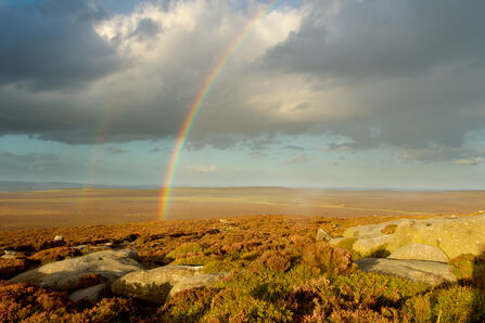 Bright rainbow stretching across a rocky moorland under dramatic dark clouds, with sunlight illuminating the landscape and a faint second rainbow visible in the distance.