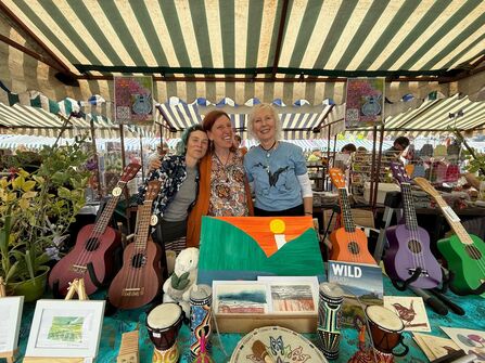 Three people standing behind a market stall displaying colourful ukuleles, drums, artwork and crafts beneath a striped canopy at an outdoor fair.