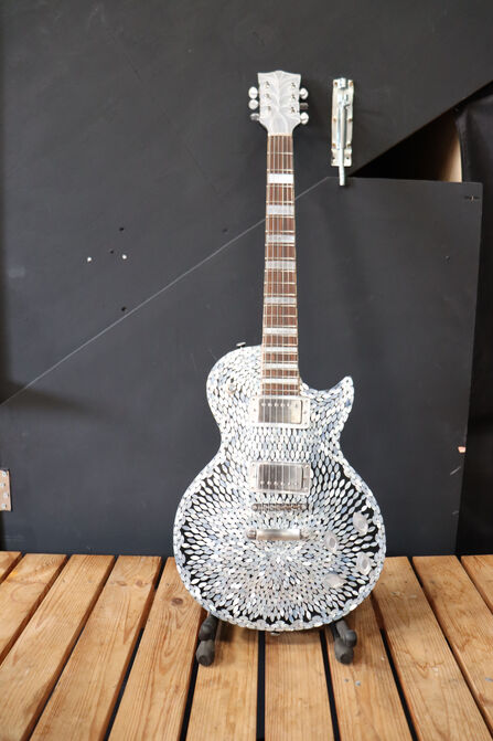 Decorative electric guitar with a iridescent pearl ovals radiating from the centre, standing on a floor stand against a dark wall.